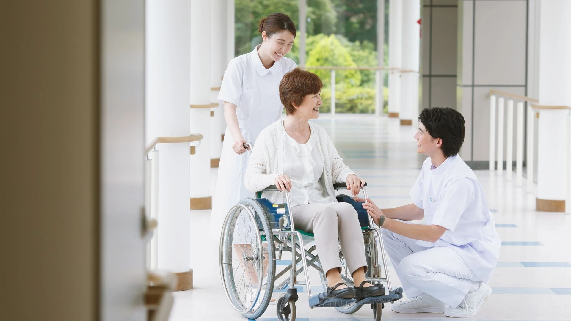 Nurse assisting senior woman in wheelchair while another nurse offers support in a bright clinic hallway.