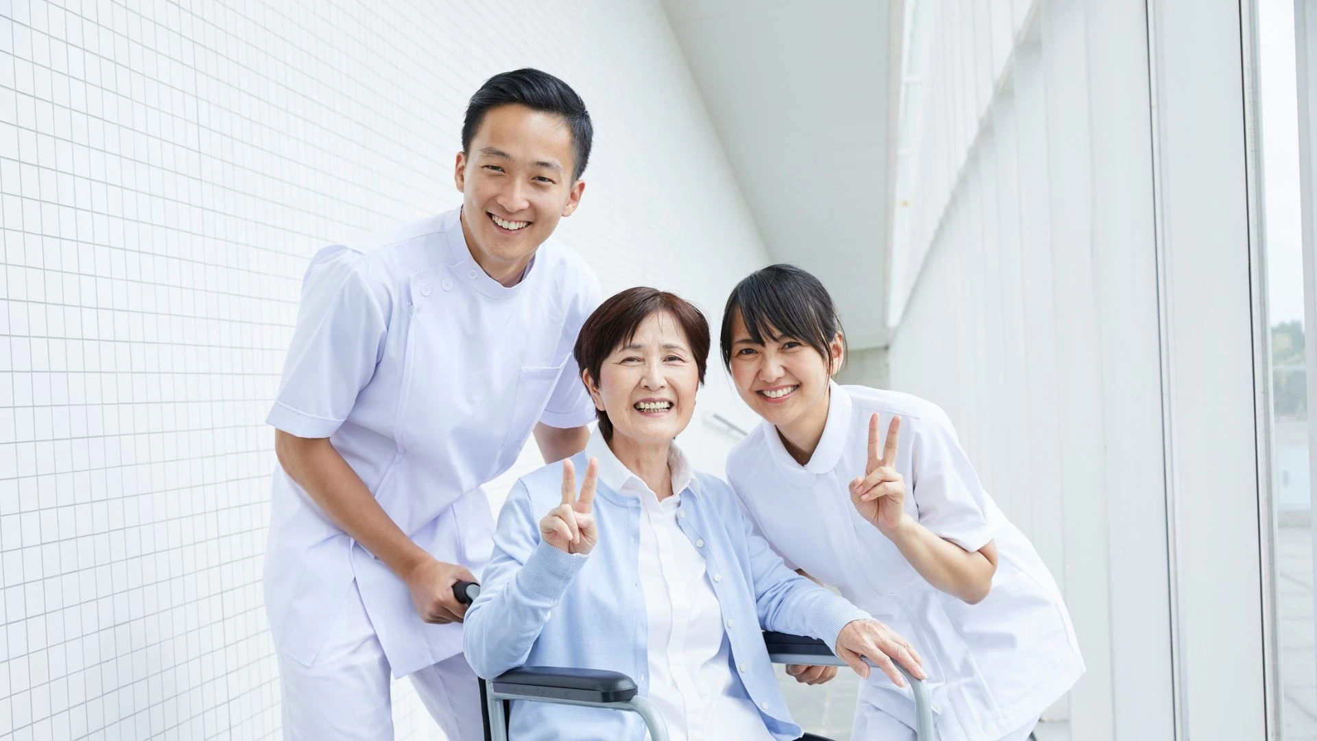 Patient in wheelchair smiling and making peace signs with two nurses in a bright corridor.